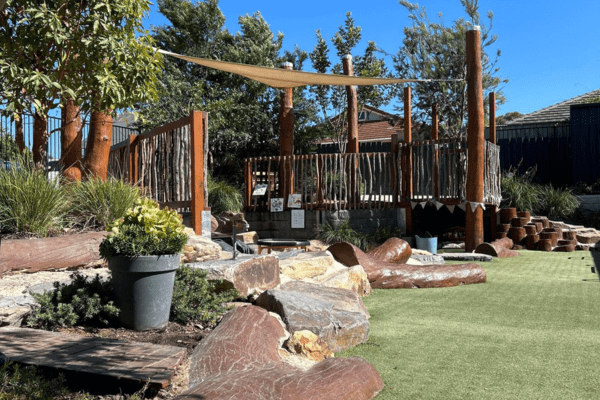 Outside play space of Curiosity Early Learning Centre featuring large timber logs and sandstone rocks for climbing, landscaped gardens with shade trees, and a large artificial turf area.