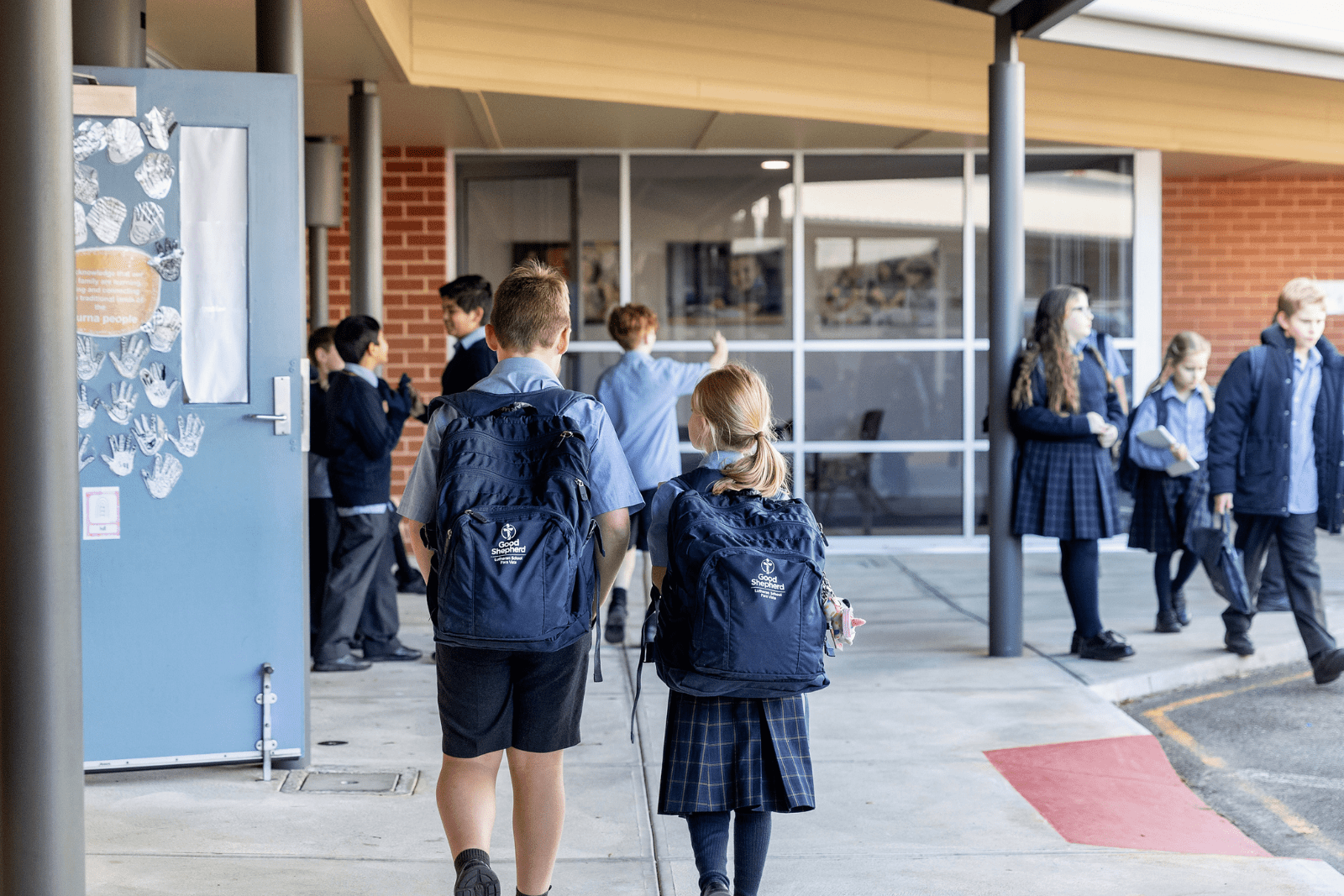 Two students are walking away from the camera. They are wearing school bags. Students are in the background walking to class.