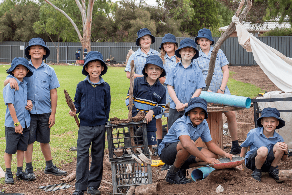 A group of students are playing outside next to the school oval. They are all wearing school uniform. They are smiling at the camera.
