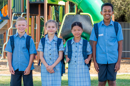 Four students in school uniform are smiling at the camera. There is a playground behind them.