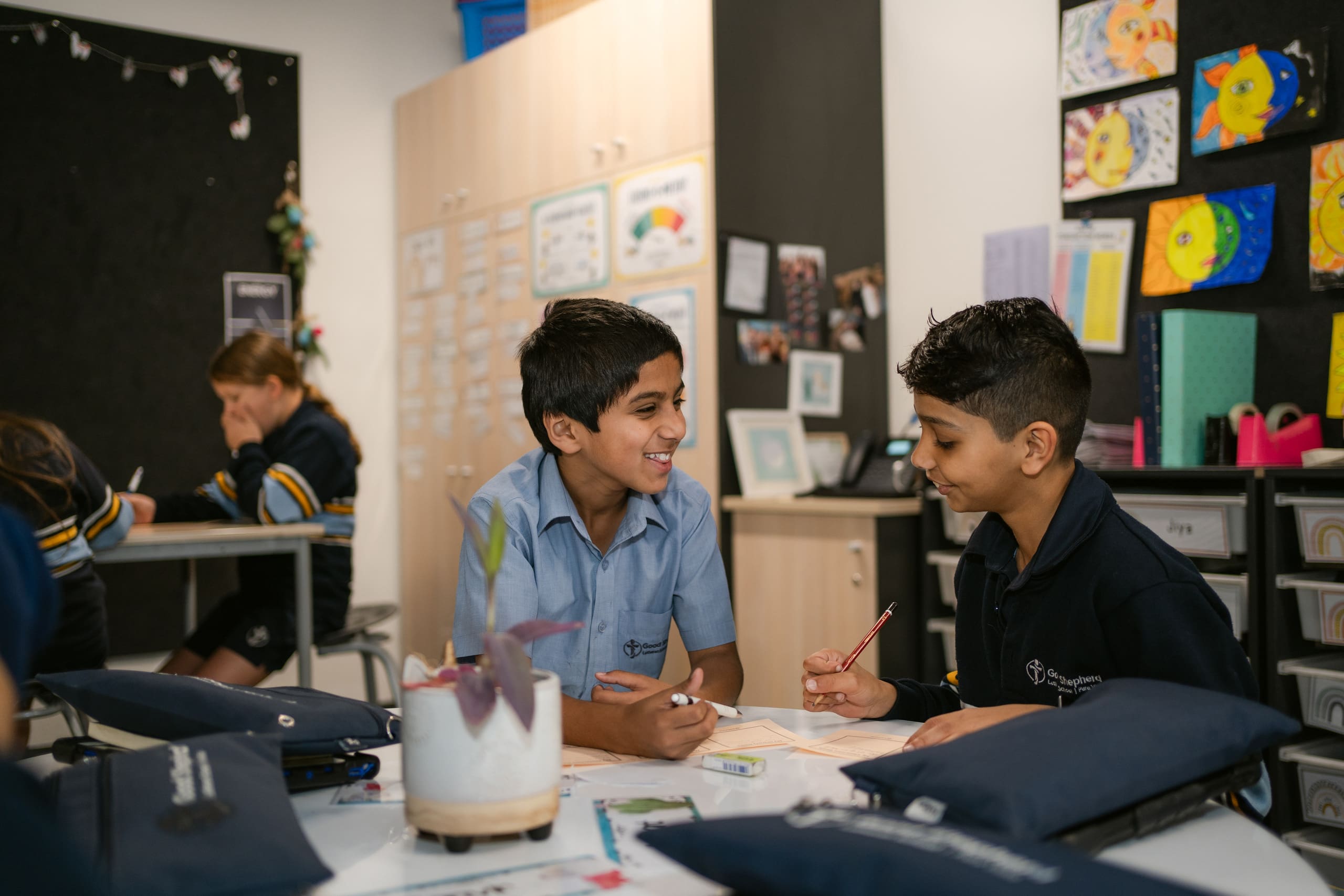 Two students are sitting at a desk in a classroom. They are smiling.