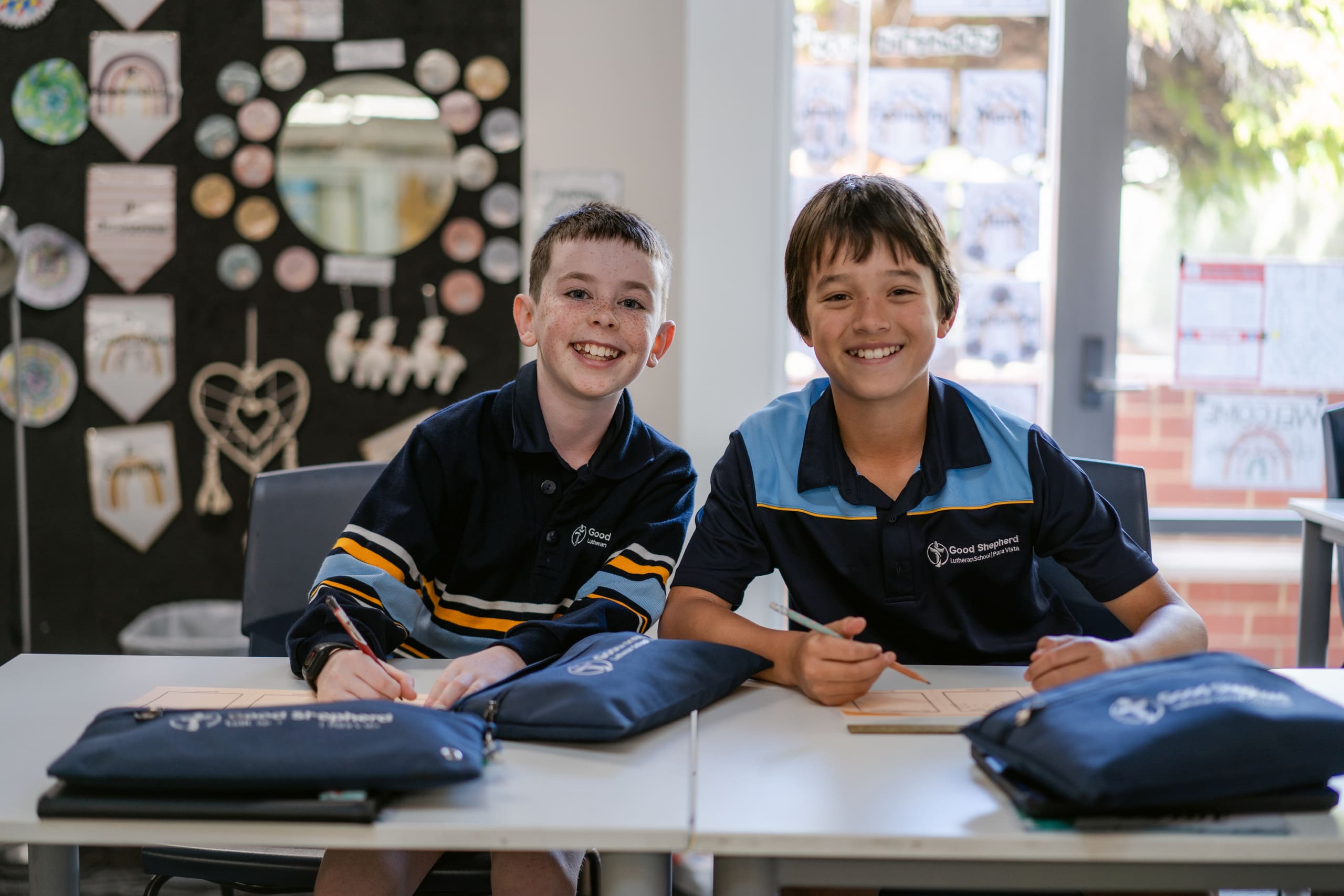 Two students are sitting at a desk in class. They are smiling at the camera.