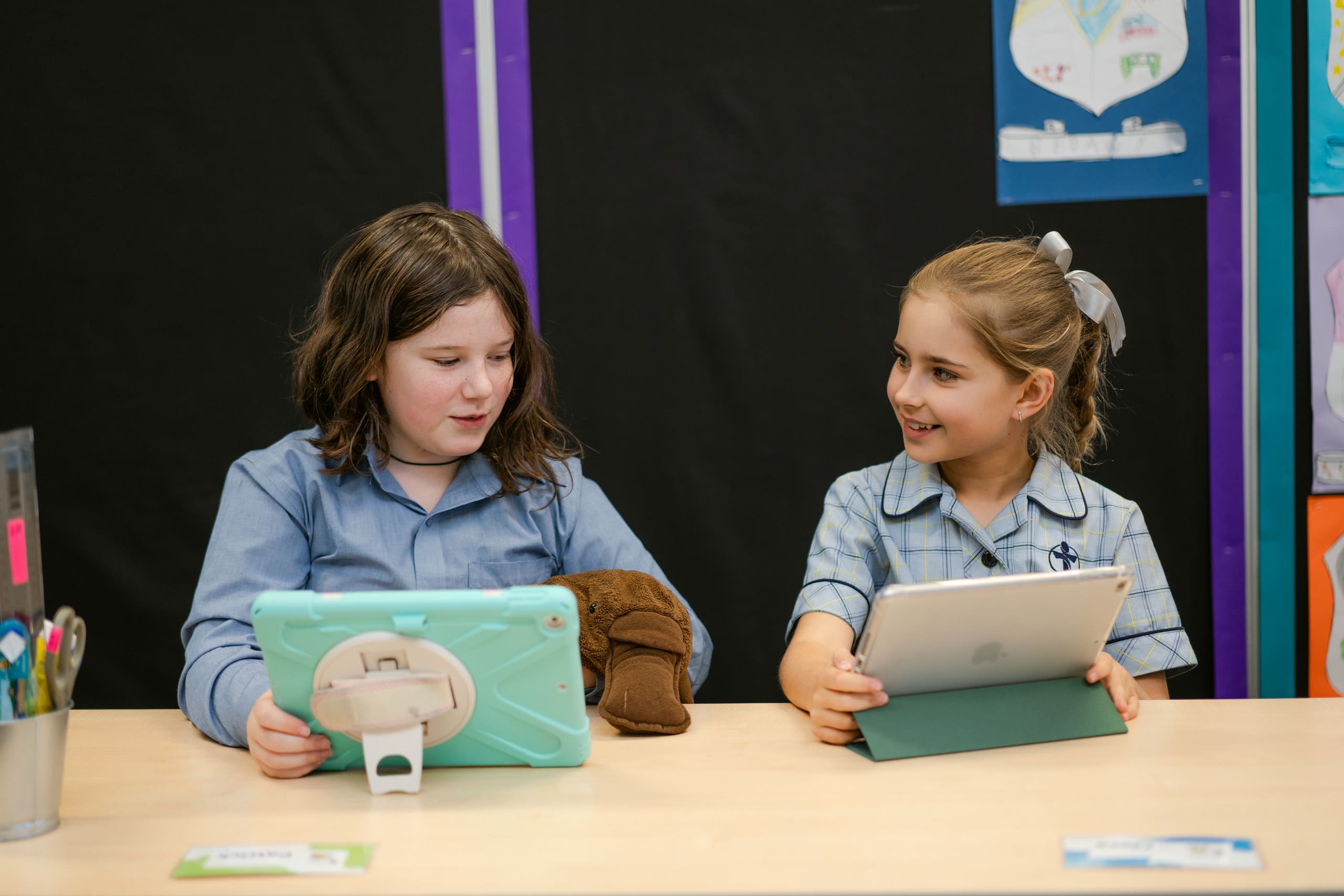 Two students at a desk using Ipads. They are smiling at each other.