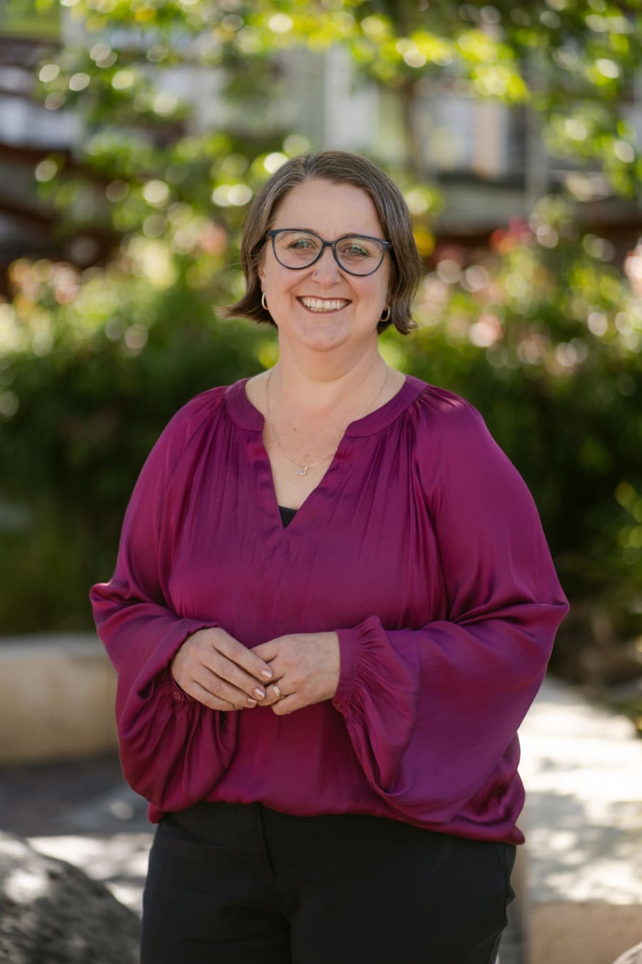 Portrait photo of Principal Evie Stevens. She is wearing a dark magenta long sleeve top and black pants. She is looking at the camera and smiling. There is a garden behind her.