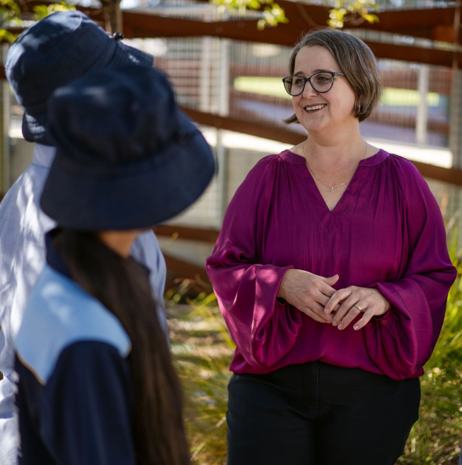 Principal of Good Shepherd Lutheran School, Evie Stevens, is talking to two students. The students have their back to the camera. Mrs Stevens is smiling at the children. She is wearing a deep pink long sleeve satin top and dark pants. There is a gard behind her.