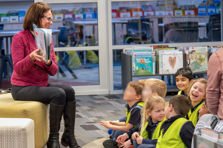 A staff member of Good Shepherd Lutheran School is reading a book to a group of young children from Curiosity ELC in the school library. The students are sitting on the floor, they are looking at each other and laughing. The staff member is holding up a book and smiling at the children.