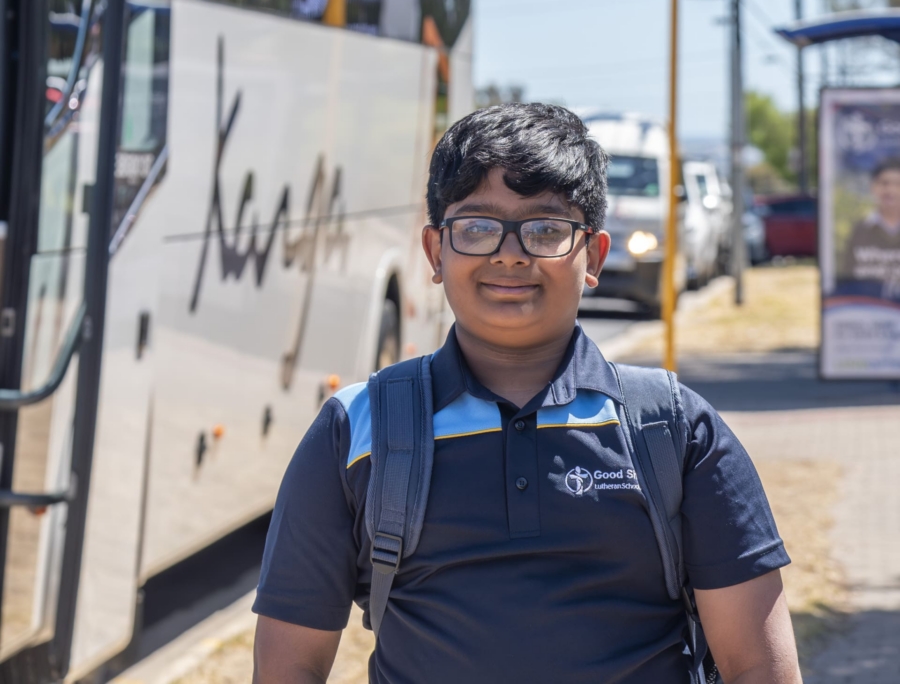 Close up portrait of a student of Good Shepherd Lutheran School. He is wearing the GSPV school uniform. He is standing in front of the school bus. There is a bus stop in the background.