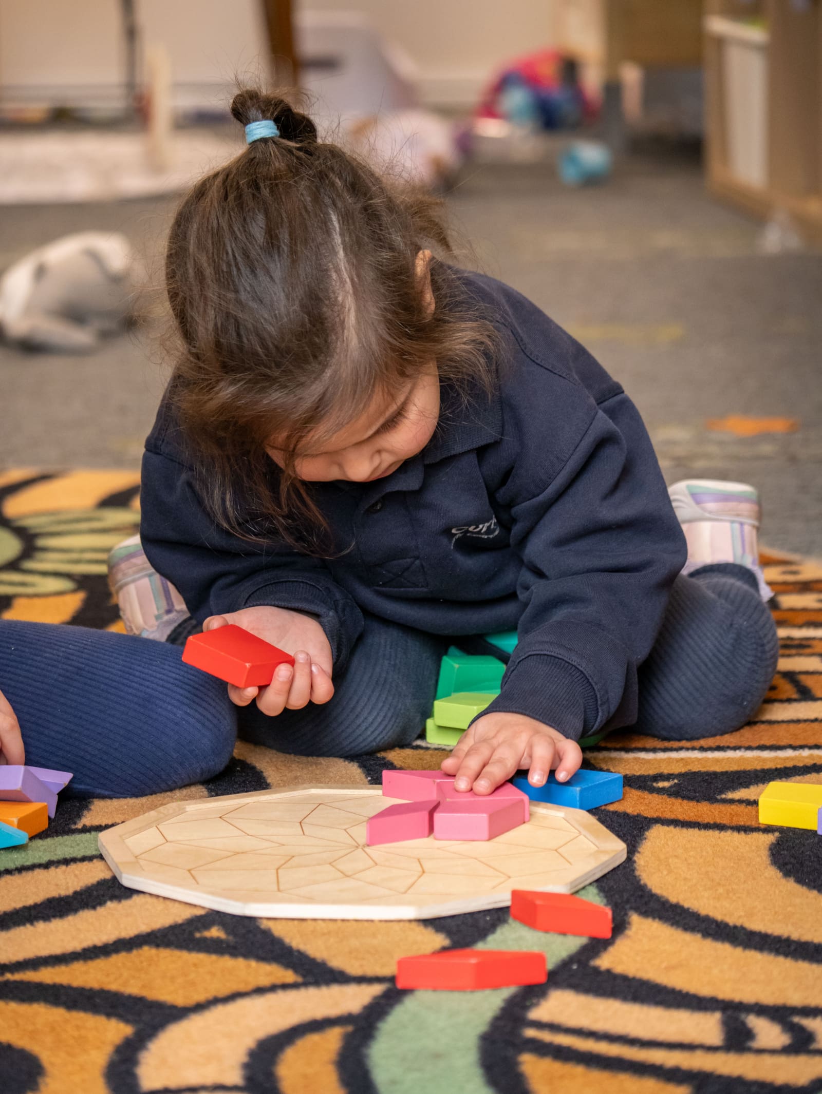 A young child, student of Curiosity Early Learning Centre, is sitting on the floor solving a colourful wooden puzzle.
