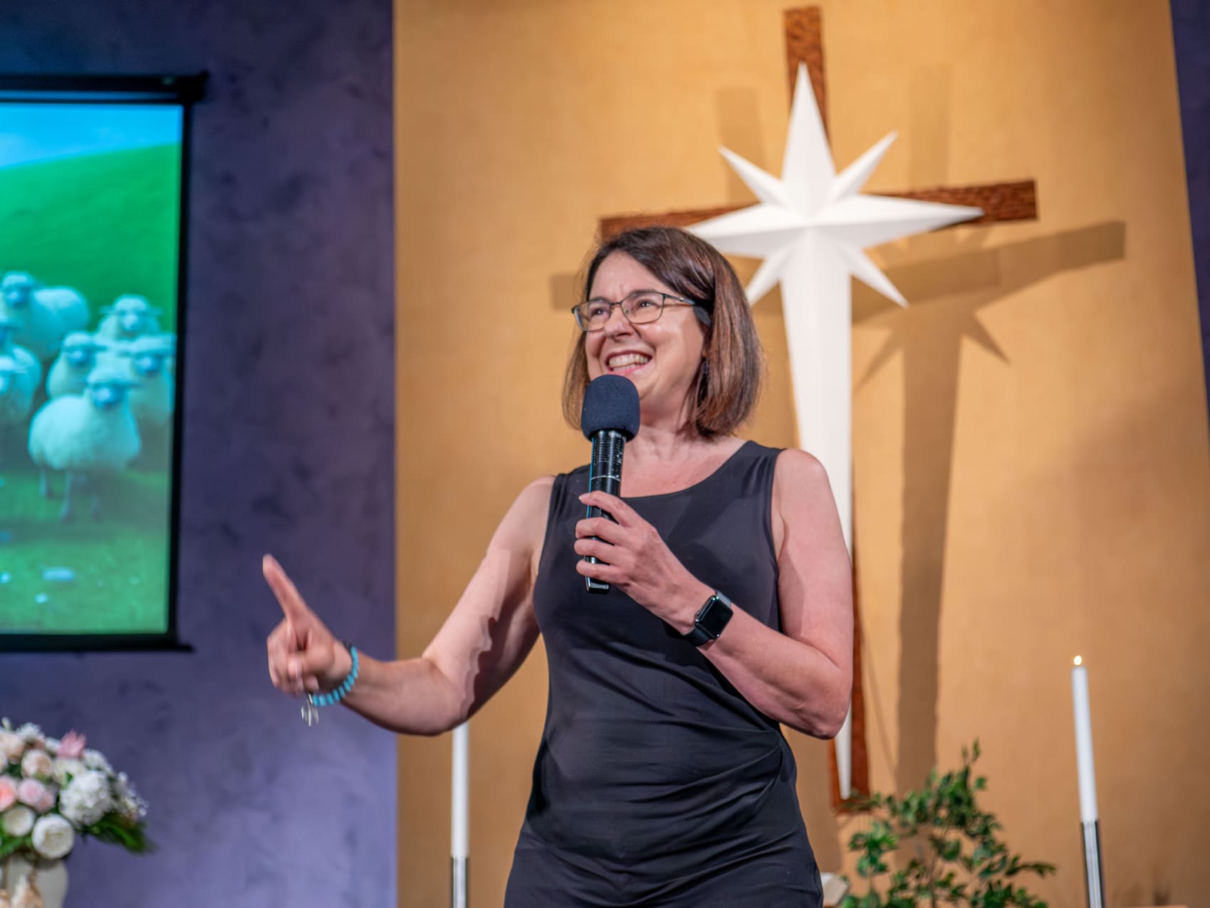 A staff member of Good Shepherd Lutheran Church and school is on stage at worship talking into a microphone. She is smiling.