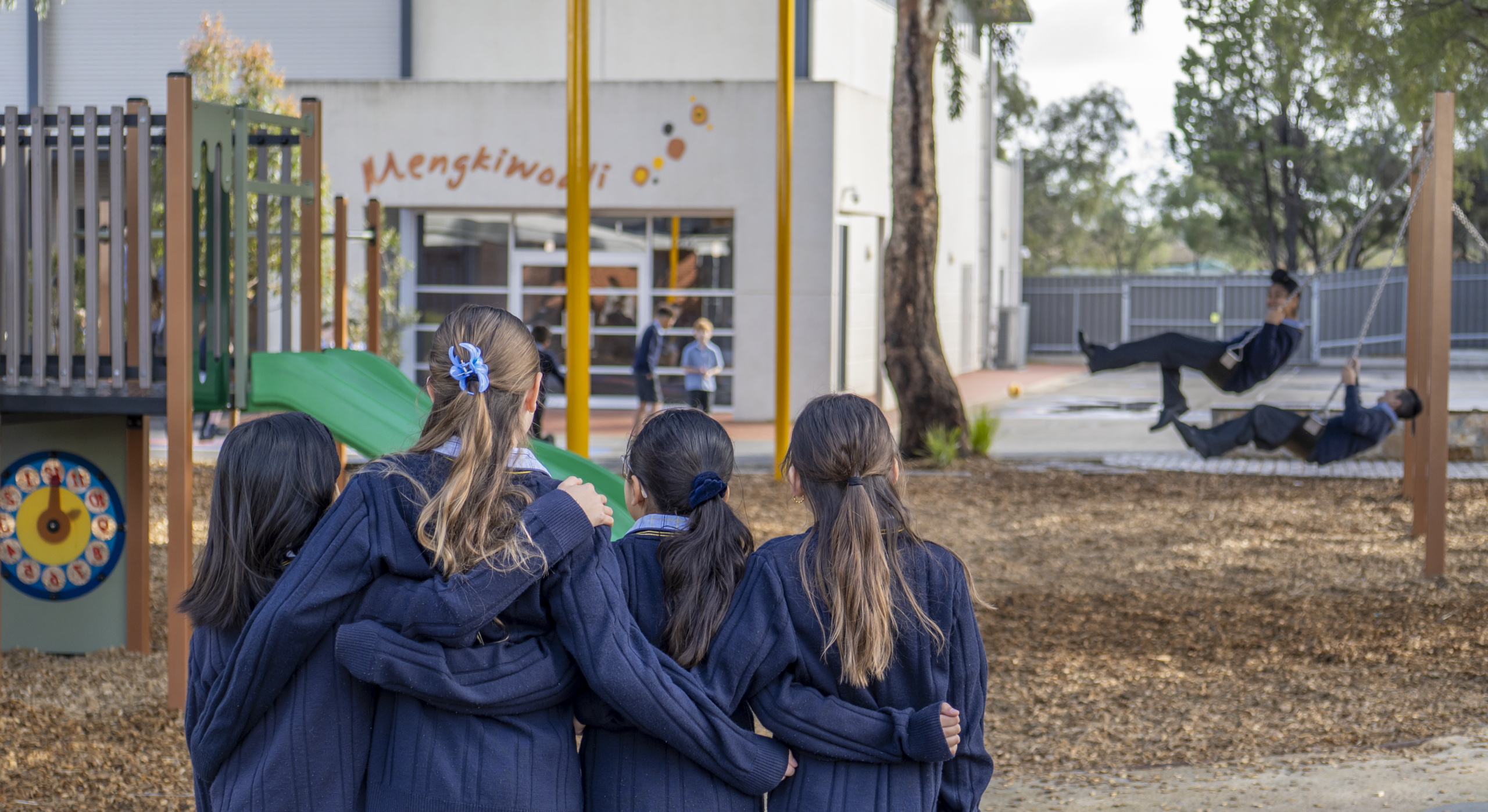 Four students of Good Shepherd Lutheran School are facing away from the camera and have their arms around each other. There are children playing on the playground in the background.